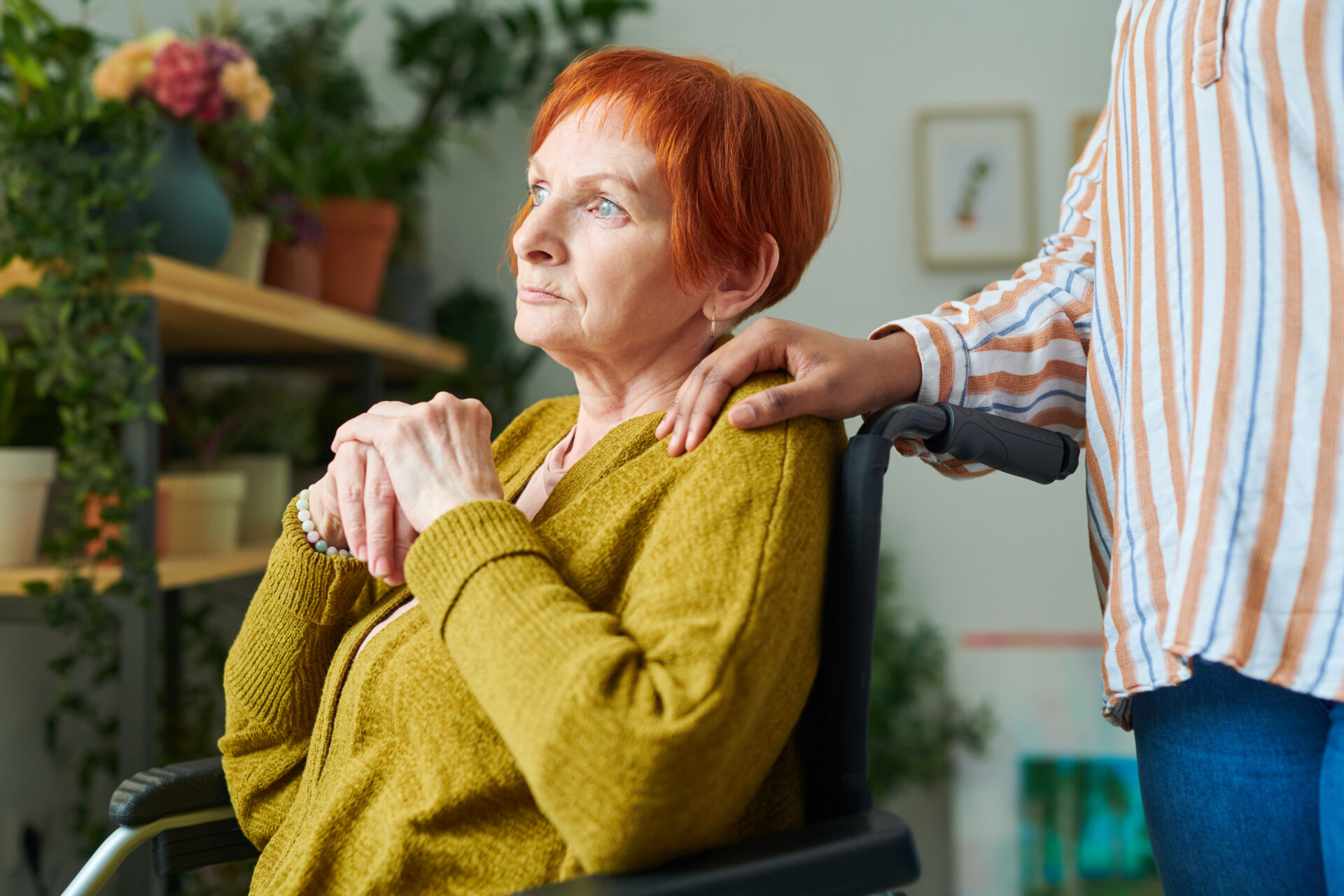 Elderly woman with red hair sitting on wheelchair and looking at window with thoughtful expression with caregiver holding her shoulder behind