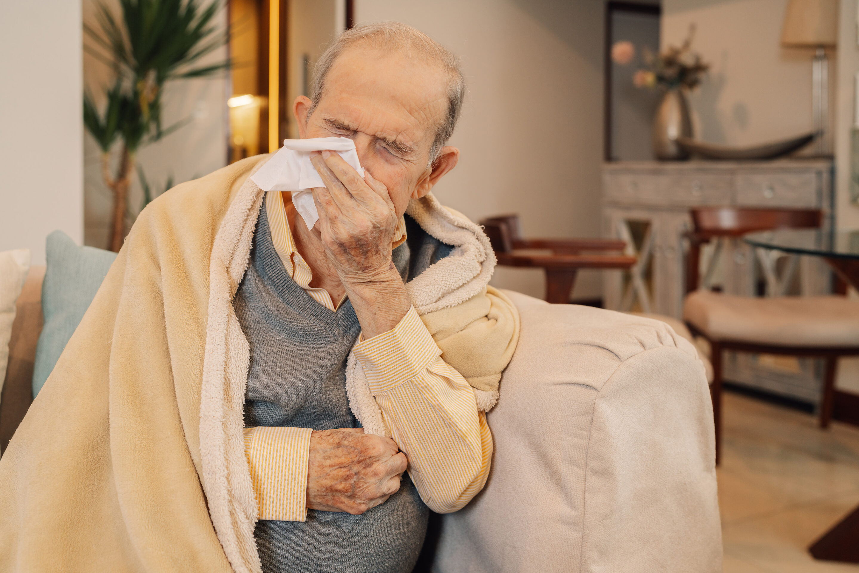 Senior man sitting on a sofa, covered with a blanket, blowing his nose while sick with the flu.
